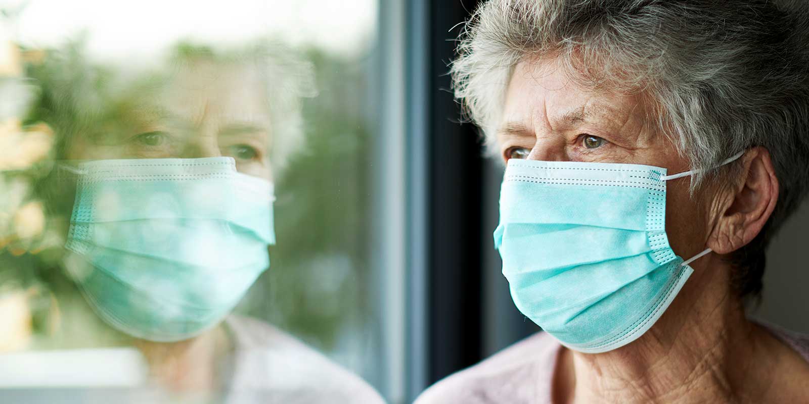 Older woman with medical mask staring out window of her home