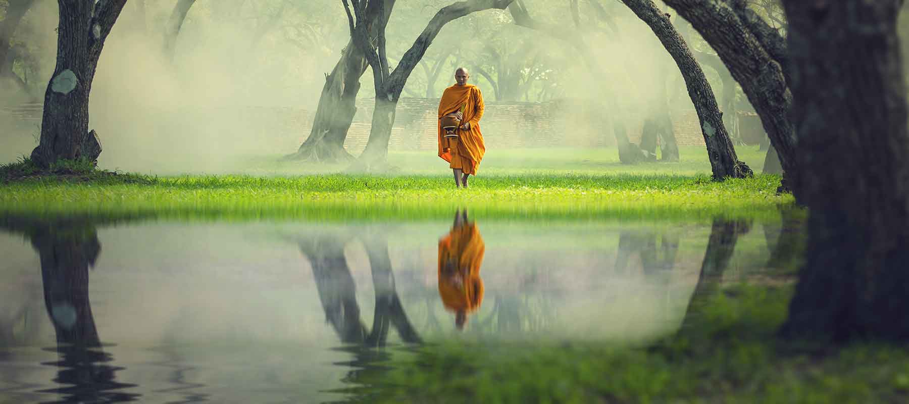 A monk walking towards water in a forest with his reflection in the water.