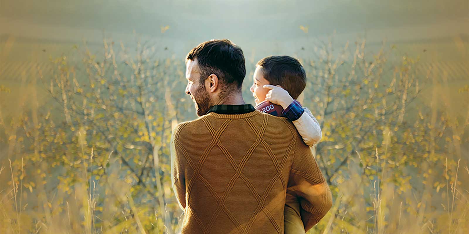 A father holding his son as they walk through a field of tall grass.