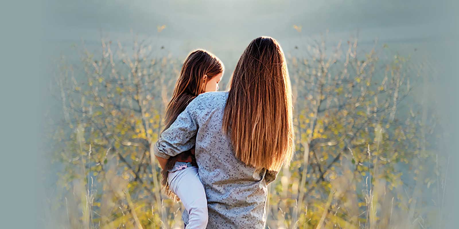 A mother holding her daughter as they walk through a field of tall prairie grass.
