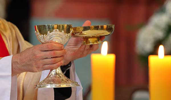 A priest holds a chalice with a candle in the background