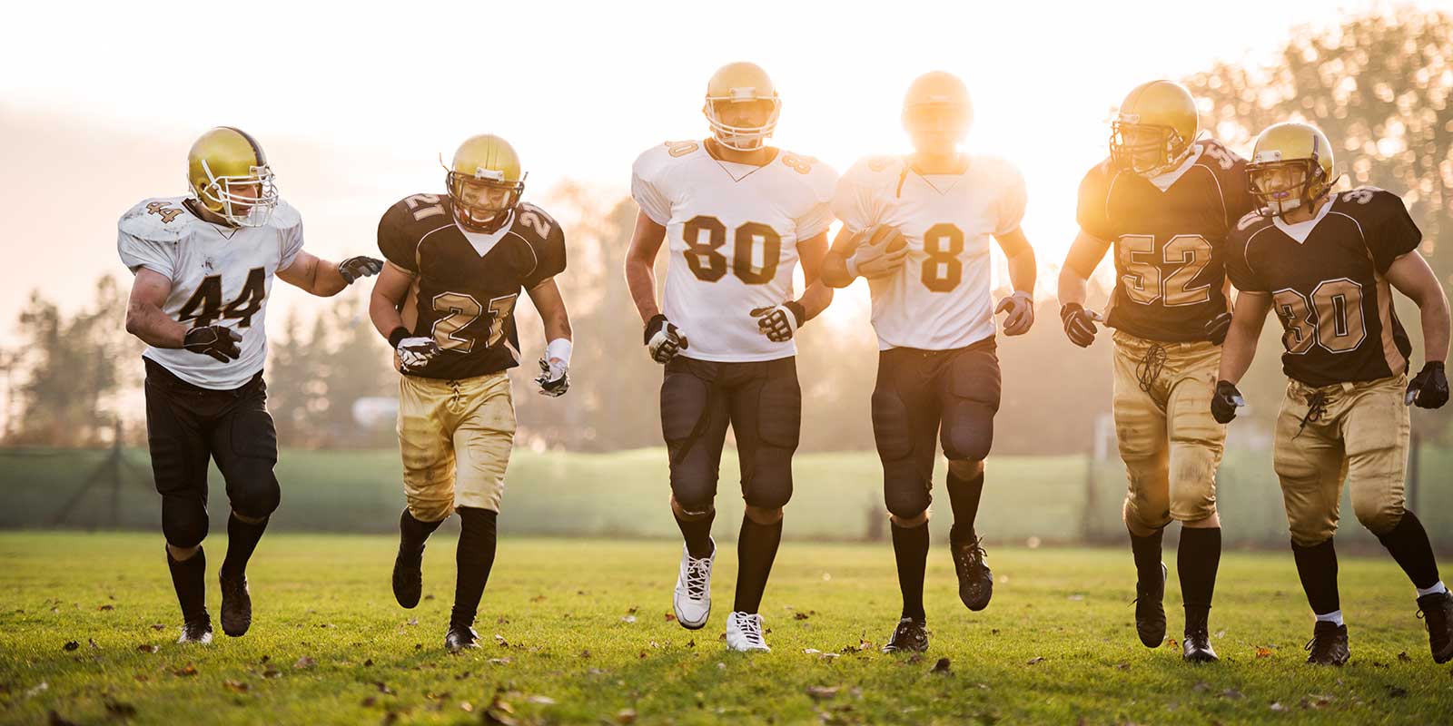Football players facing forward and jogging on a field in warm daylight.